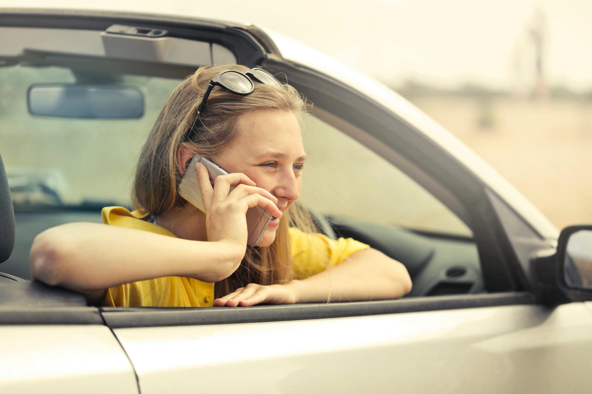 Woman smiling while talking on phone in a convertible car during a sunny day.